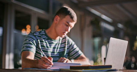 Man, college student and outdoor with laptop or writing on notebook for assignment on campus. Male person, university learner and textbook for education with research or study for exam and assessmentの写真素材