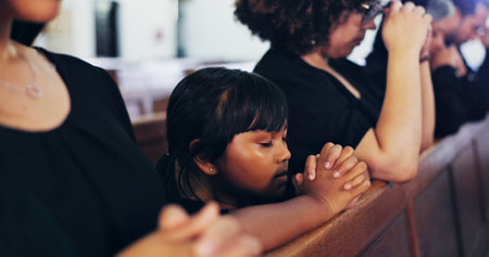 Religion, praying and kid in church with family for memorial, burial or funeral service. Worship, spiritual guidance and girl child with prayer for hope, faith or respect with gospel in chapel.の写真素材