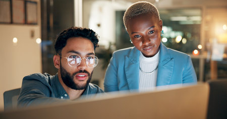 Collaboration, computer and night with employee people in office together for development or planning. Conversation, feedback or review with business man and woman at work for teamwork in eveningの写真素材