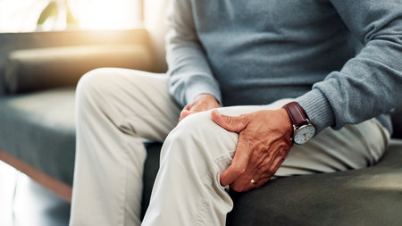 Hands, knee pain and old person on couch with injury, retirement and health issue with muscle tension at home. Fibromyalgia, arthritis or brittle bones with legs, inflammation and pressure in houseの写真素材