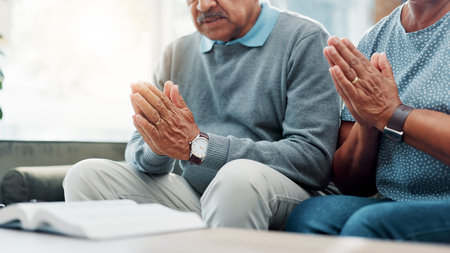Hands, senior couple and bible with prayer on sofa for religious worship, spiritual praise or faith in Jesus. Old people, grace or holy gospel in home for scripture meditation, thankful or connectionの写真素材
