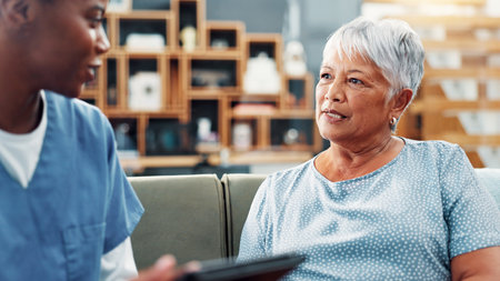 Senior woman, nurse and conversation in home for healthcare, medical support and wellness. Retirement, patient and talking with caregiver in living room for help, checkup and insurance with tabletの写真素材