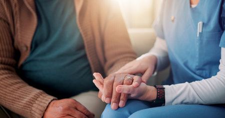 Sofa, caregiver and senior patient with holding hands for support, empathy and console for medical news. Home, nurse and old person with consultation for wellness, compassion and flare in closeupの写真素材
