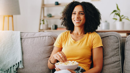 Happy woman, relax and watching tv with bowl of popcorn on sofa for online entertainment at home. Female person, user or eating with smile, snack or food for streaming show, movie or series at houseの写真素材