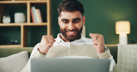 Man, fist pump and laptop on couch with celebration, excited and winning with online giveaway in home. Person, computer and happy with goal, success and profit with investment in living room at houseの写真素材