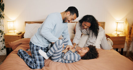 Parents, child and play on bed in family home for bonding, tickle or energy at night. Interracial dad, kid and funny game with mother in bedroom for love, care or laugh together with happy foster boyの写真素材