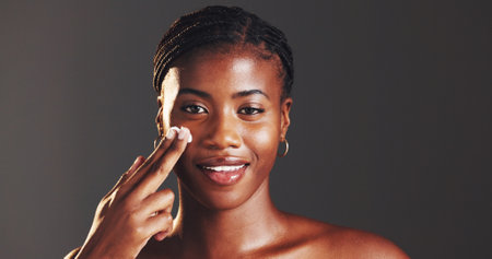 Portrait, beauty and woman with cream, skincare and smile for smooth skin with routine and moisturizing. Gray background, hydration and black person with lotion for treatment, studio and groomingの写真素材
