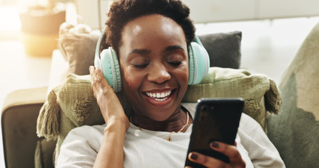 Headphones, black woman and relax with phone on sofa for wellness podcast, stress relief and music playlist. Home, person and smartphone with eyes closed for calm sound, zen audio and weekend breakの写真素材