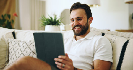 Home, smile and man on couch, tablet and internet with online video for humor, joke and social media. Apartment, tech and happy person on sofa, meme and laughing with digital app for entertainmentの写真素材