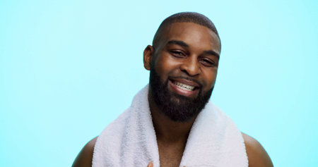 Black man, portrait and smile with towel in studio for natural beauty, beard and wellness on blue background. Shower, cosmetic or happy male person with facial hair, grooming or space for cleaningの写真素材