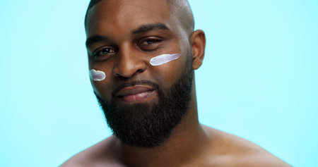 Portrait, cream and black man in studio, skincare and dermatology on blue background. African person, space and model with shine, creme and lotion with moisturizer, beauty and facial spf with glowの写真素材
