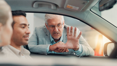Drive, salesman and couple with talking in car dealership, transport model and rental service. Showroom, people and mature seller with pitch for vehicle features, assistance and discussion by flareの写真素材