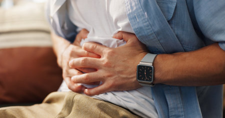 Person, hands and stomach ache with gastro infection, indigestion or constipation on sofa at home. Closeup, patient or sore belly with tummy for food poisoning, diarrhea or abdominal muscle strainの写真素材