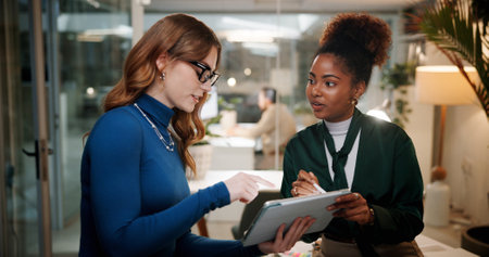 Business women, night and discussion with tablet for design, project or schedule at office. Female people, employees or colleagues working late with technology for tasks, ideas or agenda at workplaceの写真素材