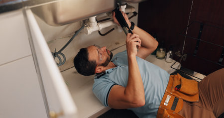 Man, plumber and repair in kitchen, pipe and sink with search for leak, problem solving and assessment in home. Above, handyman and tools for job, drain and solution for maintenance at apartmentの写真素材
