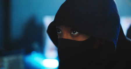 Serious, hacker and person with mask, night and mysterious for cyber attack in office, scam and privacy. Anonymous, criminal and security breach for ransomware, hacking and cover for face or fraudの写真素材
