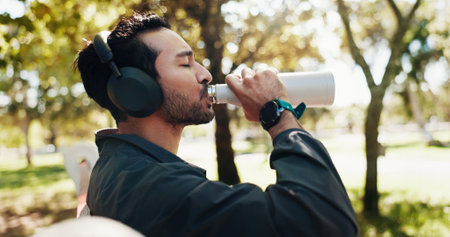 Break, drinking water and music with man in park for exercise, fitness or running routine. Headphones, hydration and streaming with athlete or runner outdoor in nature for recovery, relax and restの写真素材