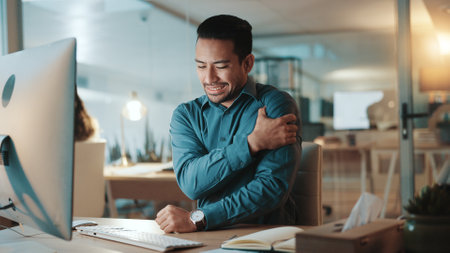 Frustrated, businessman and night with shoulder pain on computer for muscle tension at office. Man, employee or working late with sore arm, ache or injury for discomfort or bad posture at workplaceの写真素材