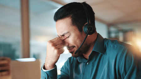 Night, call center and man in office, headache and burnout with deadline, pressure and fatigue. Evening, person and consultant with migraine, tired and tension with pain, stress and customer serviceの写真素材