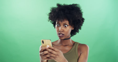 Phone, shocked and text message with black woman in studio for communication of bad news or update. Anger, app and social media with frustrated African person on green background for notificationの写真素材