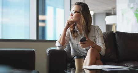 Phone, thinking and waiting with business woman in office lobby for job opportunity or vacancy. App, resume and vision with candidate on sofa in workplace for hiring, interview or recruitmentの写真素材