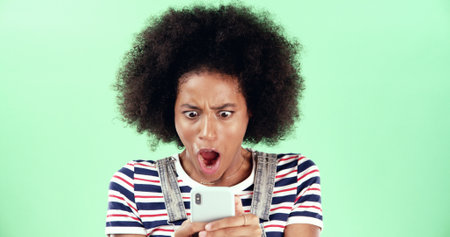 Black woman, smartphone and shock with open mouth in studio, what or fake news with alert for online message. Surprise announcement, alarm or wow expression with WTF on mobile app on green backgroundの写真素材
