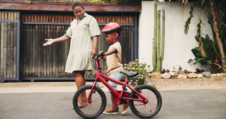 Support, learning and boy on bicycle with mom on street of neighborhood for child development. African woman, love or black kid with trust, help or single mother teaching son how to ride bike outsideの写真素材