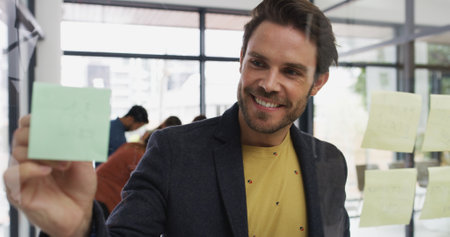 Glass wall, businessman and smile at sticky note for writing, strategy and brainstorming in agency. Office, male person and creative writer with moodboard for information, solution and startup goalの写真素材