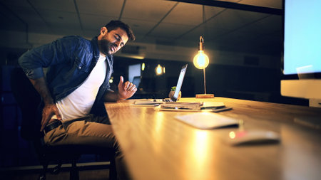 Laptop, back pain and programmer at night for research, web design deadline or office project. Creative worker, administrator and fatigue for overtime, update or burnout for website article onlineの写真素材