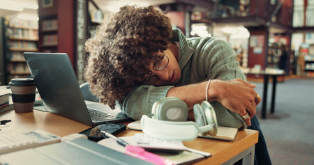 Tired, man and student sleeping in library with burnout for studying with university test. Fatigue, education and person with afternoon nap for resting at desk laptop at college campus for exhaustionの写真素材