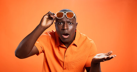 Surprised, man and question for news, studio and facial expression for revelation, what and fashion. Orange background, shocked and black person with sunglasses, portrait and mockup space for gossipの写真素材