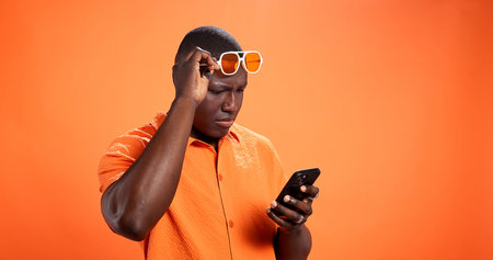 Black man, phone and confused in studio with chat, thinking and reading post on orange background....