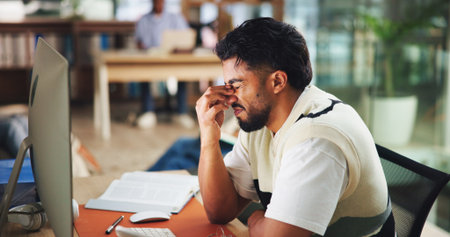 Frustrated, headache and man at university in library for learning, education and studying. College, school and student with notes, books and pc for project with eye strain, burnout and stressの写真素材