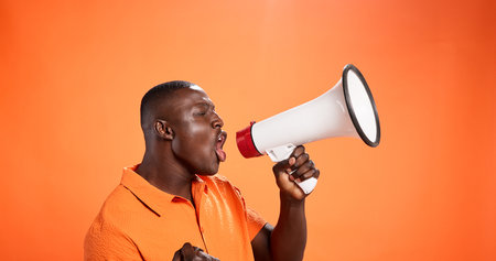 Black man, megaphone and voice in studio for announcement, broadcast or opportunity. Join us, mockup space or African speaker screaming with noise, giveaway speech or attention on orange backgroundの写真素材