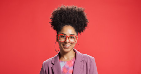 Portrait, smile and woman for fashion in studio with earrings, glasses and trendy clothes. Happy, African female person and attitude with jewellery, stylish outfit and accessories on red backgroundの写真素材