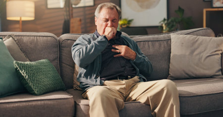 Senior, sick man and couch with chest pain on sofa for cardiac arrest or bronchitis at home. Elderly, male person or patient with illness, virus or viral infection for lung disease or heart attackの写真素材