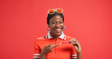 Portrait, lip gloss and black woman in studio, pointing and promotion on red background. African person, makeup and model with cosmetics, announcement and advertising space with beauty and smileの写真素材