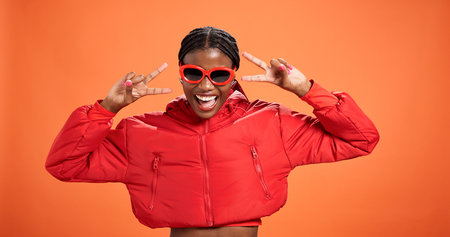 Sunglasses, excited and black woman with peace sign for fashion, style or trendy. Happy African model, studio or v hand gesture for victory symbol, emoji or cool influencer laugh on orange backgroundの写真素材