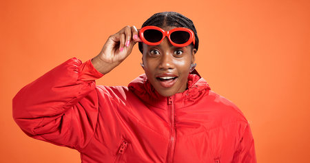 Portrait, surprise and black woman in studio, sunglasses and announcement on orange background. Facial expression, shocked and African person with shades, discount deal and crazy sale with accessoryの写真素材