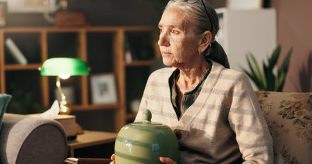 Sad, old woman and thinking with urn in home for mourning, grief and loneliness in retirement. Loss,...