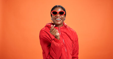 Heart, fashion and portrait of woman in studio for love, support and trendy style on orange background. Happy, smile and person with emoji, gesture and fingers for gratitude, thank you and confidenceの写真素材