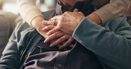Comfort, hands and elderly couple in home, bonding and affection for relationship in lounge and hug. Retirement, care and love in living room, senior people and embrace for support in house or trustの写真素材