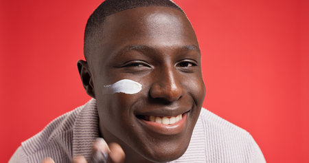 Apply, smile or portrait of black man with face cream for skincare, glow or facial beauty. Space, red background or happy African male model in studio for lotion, sunscreen cosmetics or moisturiserの写真素材