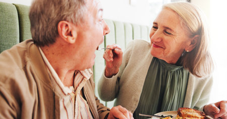 Eating, feed or senior couple in cafe on date together in retirement for love, breakfast or food. Share, taste or elderly people in restaurant with lunch and laugh at diner for romantic anniversaryの写真素材