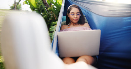 Woman, laptop and rest in hammock outdoor for research, reading email and proofreading article. Journalist, remote work and pc for story planning, post schedule and information with news publicationの写真素材