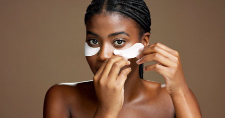 Black woman, apply and eye mask in studio for skincare, portrait or reduce puffiness. Model, brown...