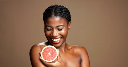 Beauty, happy and black woman with grapefruit in studio for natural, organic or facial glow routine. Dermatology, skincare and African person with citrus for face brightening by brown background.の写真素材