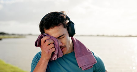 Tired man, sweat or fitness with towel or headphones at lake for intense workout, break or recovery. Active, male person or dry with cloth for physical activity, exercise or outdoor training at riverの写真素材