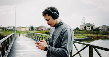 Headphones, athlete or man on bridge with phone for workout, tracking exercise or song. Lake, outdoor or runner on mobile app in city for running progress, music playlist and fitness podcast audioの写真素材