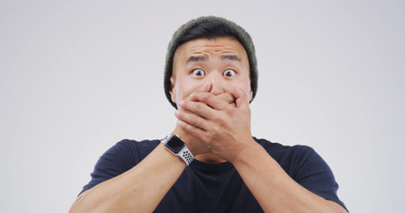 Scared, shocked and portrait of man in studio with omg, wtf or wow reaction for news. Emotion, surprise and Asian male person with hands covering mouth for rumor or announcement by white background.の写真素材